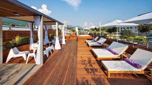 A wooden deck with white lounge chairs and tables, a covered seating area, and a view of buildings and greenery in the background. - Perla, Resort & Entertainment (Nova Gorica)