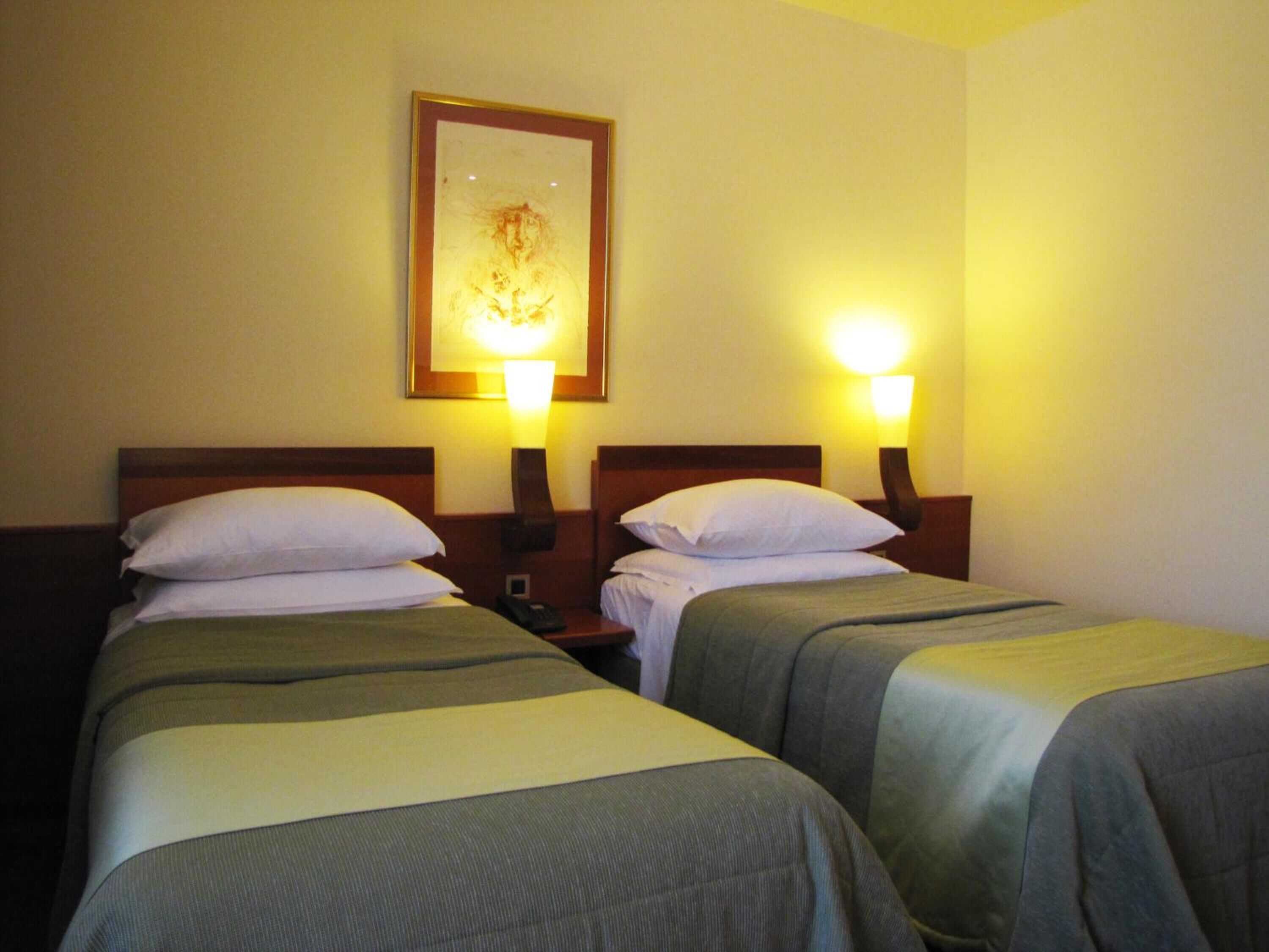 A hotel room with two single beds, each with a pillow and a bedside table with a lamp.