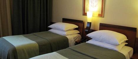 Een hotelkamer met twee eenpersoonsbedden, elk met een kussen en een deken. Daartussen staat een nachtkastje met een lamp.