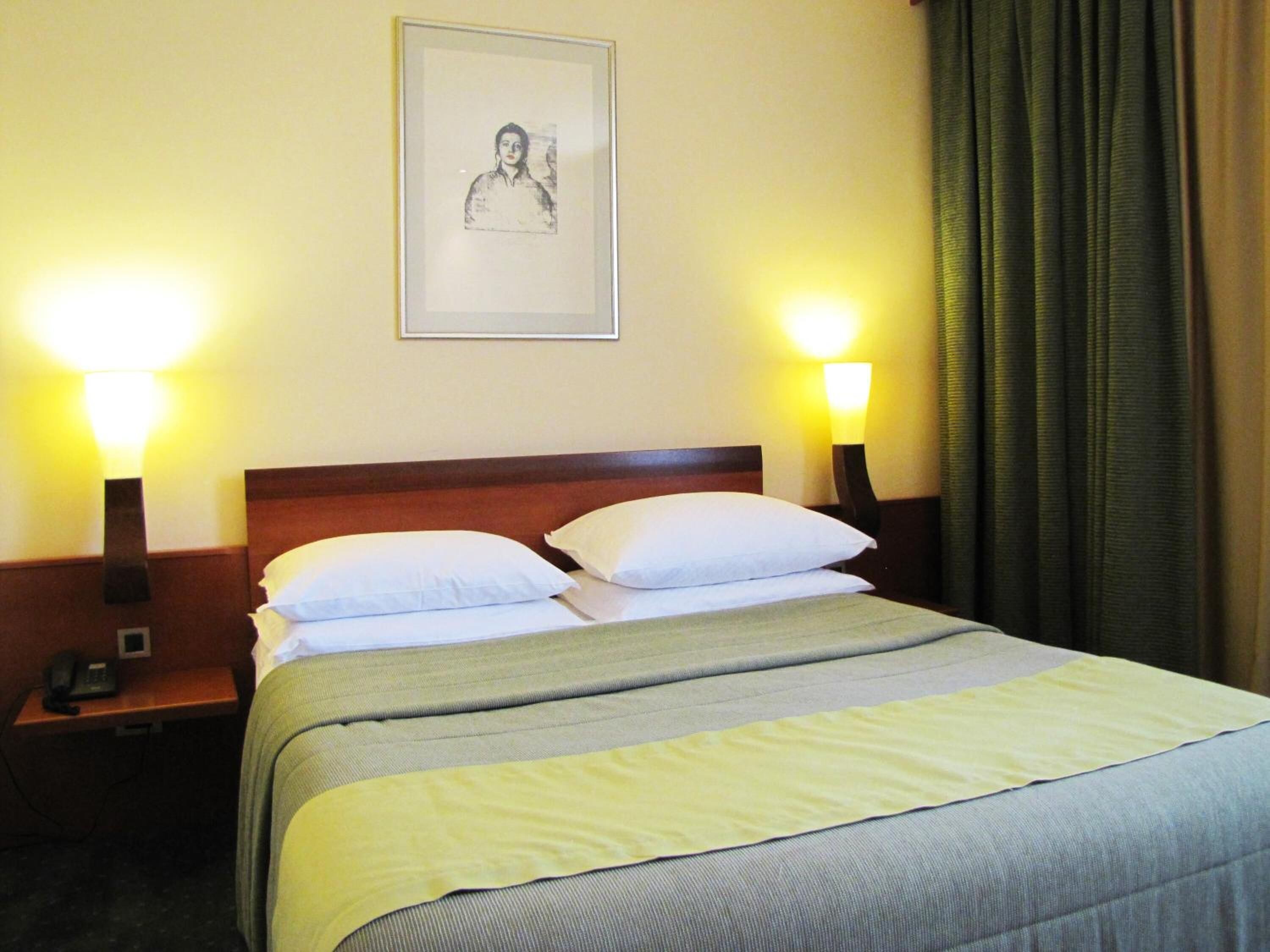 A hotel room with a bed, two bedside lamps, a framed picture on the wall, and a telephone on the nightstand.