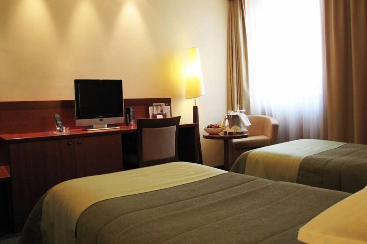A hotel room with two beds, a desk with a computer, a chair, a lamp, and a window with curtains.