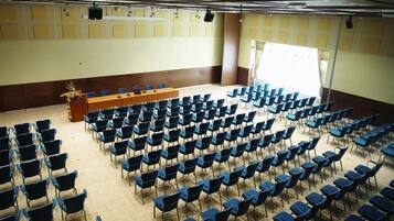 Une salle de conférence avec des rangées de chaises bleues, un pupitre en bois et une grande fenêtre.