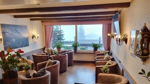 A cozy lounge area with cushioned seating, potted plants, and a large window with a view of trees and a building. - Hotel Bernerhof Grindelwald (Grindelwald)
