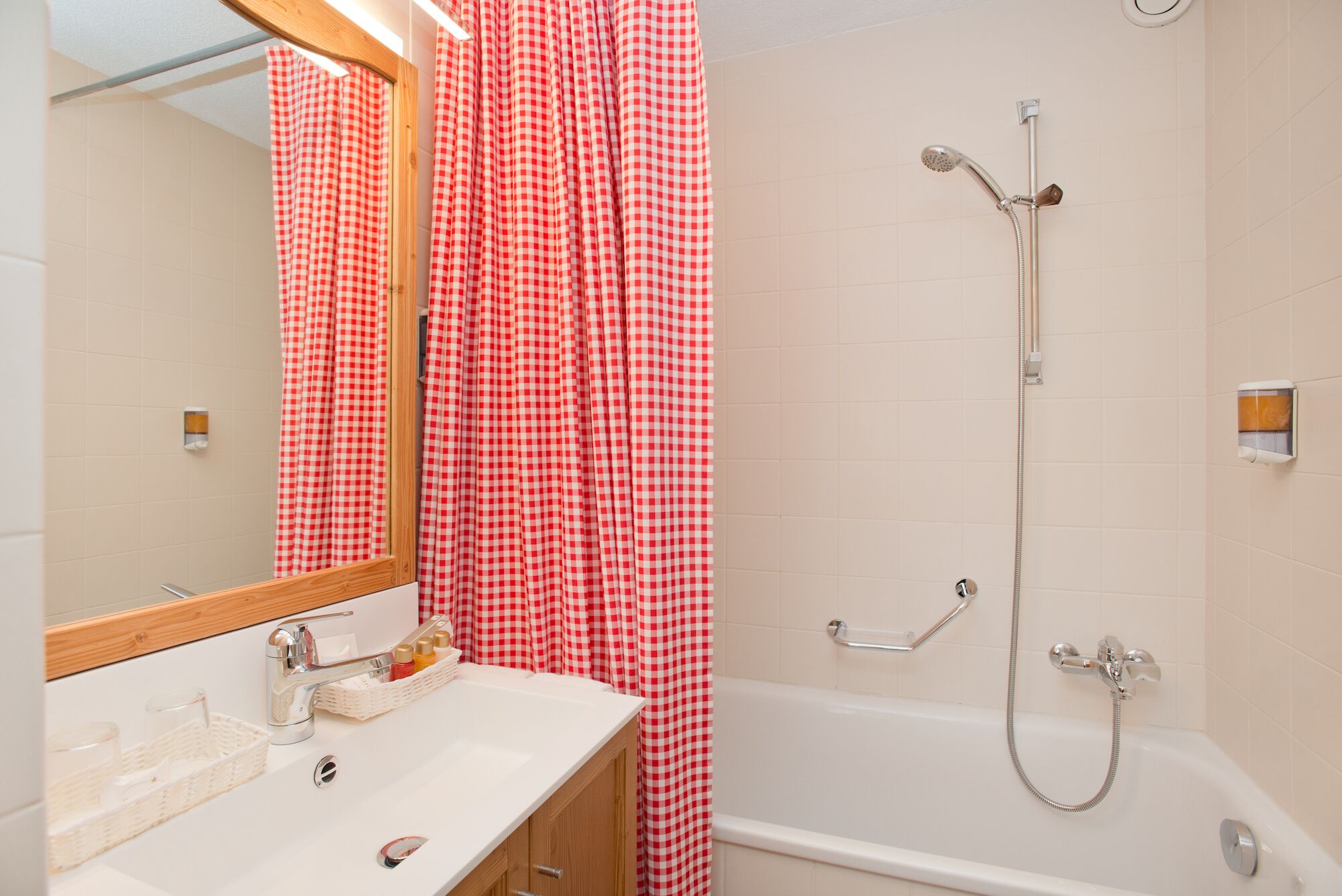 A bathroom with a white sink, wooden vanity, a mirror, a shower with a handheld showerhead, and a red and white checkered shower curtain.
