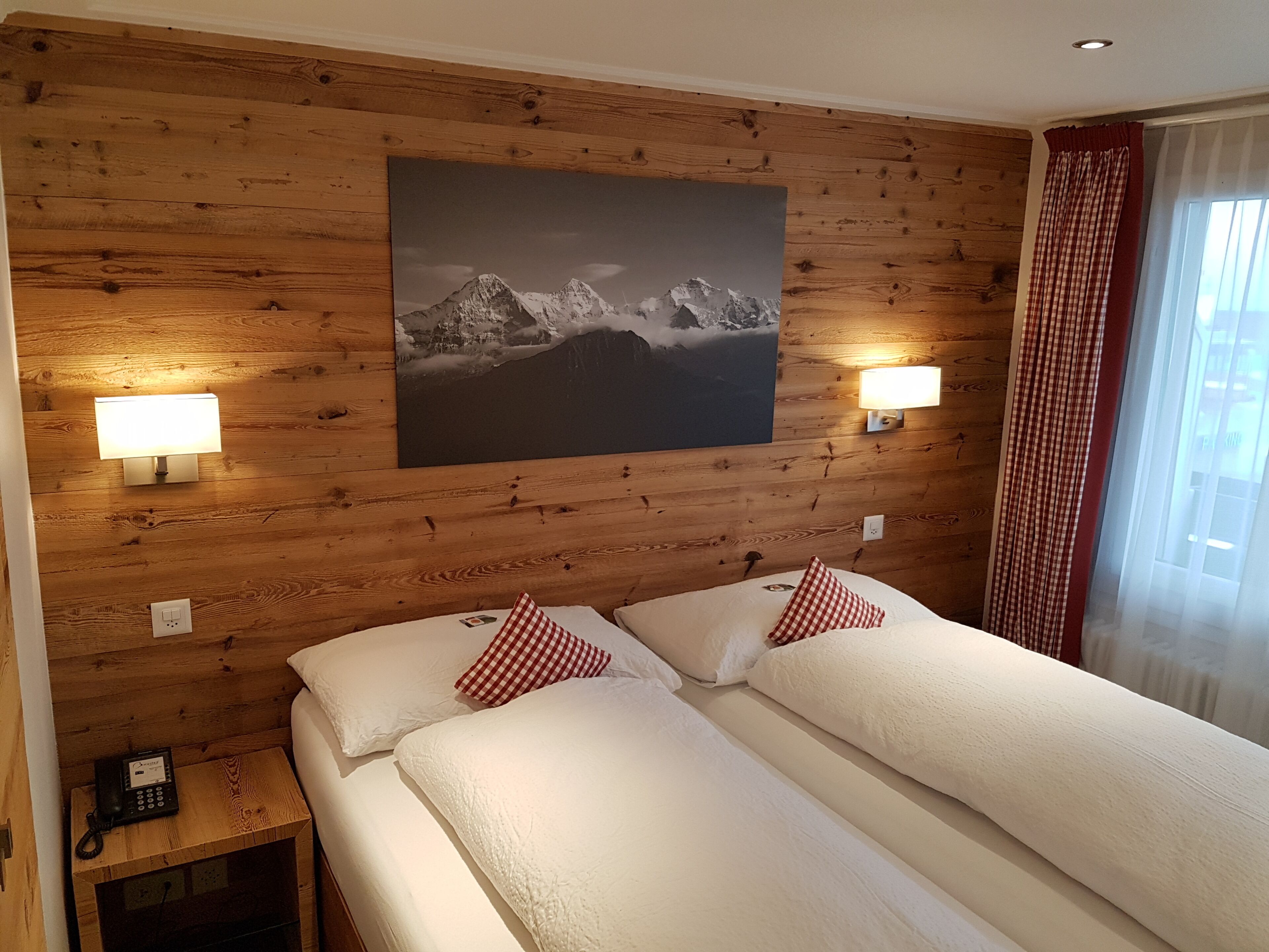 A hotel room with wooden panel walls, two beds with white bedding and red checkered pillows, a painting of a mountain landscape, and two wall-mounted lamps.