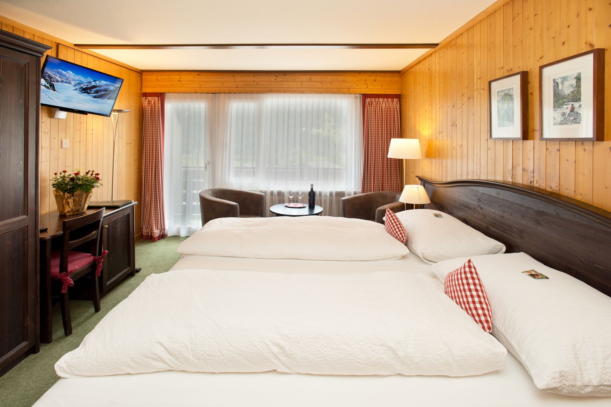 A hotel room with two beds, a TV, a desk with a chair, a small table with a bottle and glasses, and a window with curtains.
