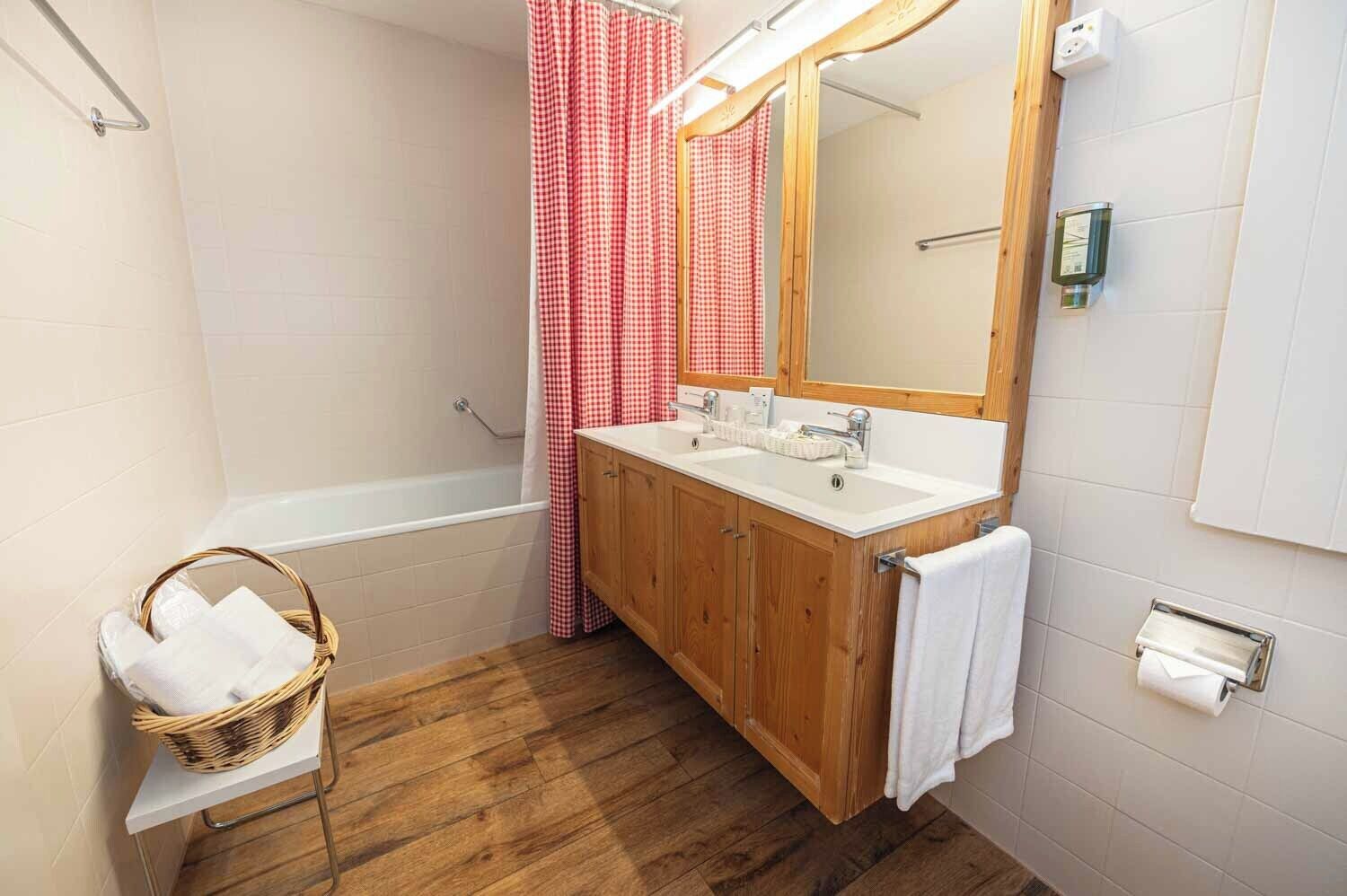 A bathroom with a wooden vanity, two sinks, a large mirror, a shower with a red and white checkered curtain, a wooden floor, and a basket with towels.