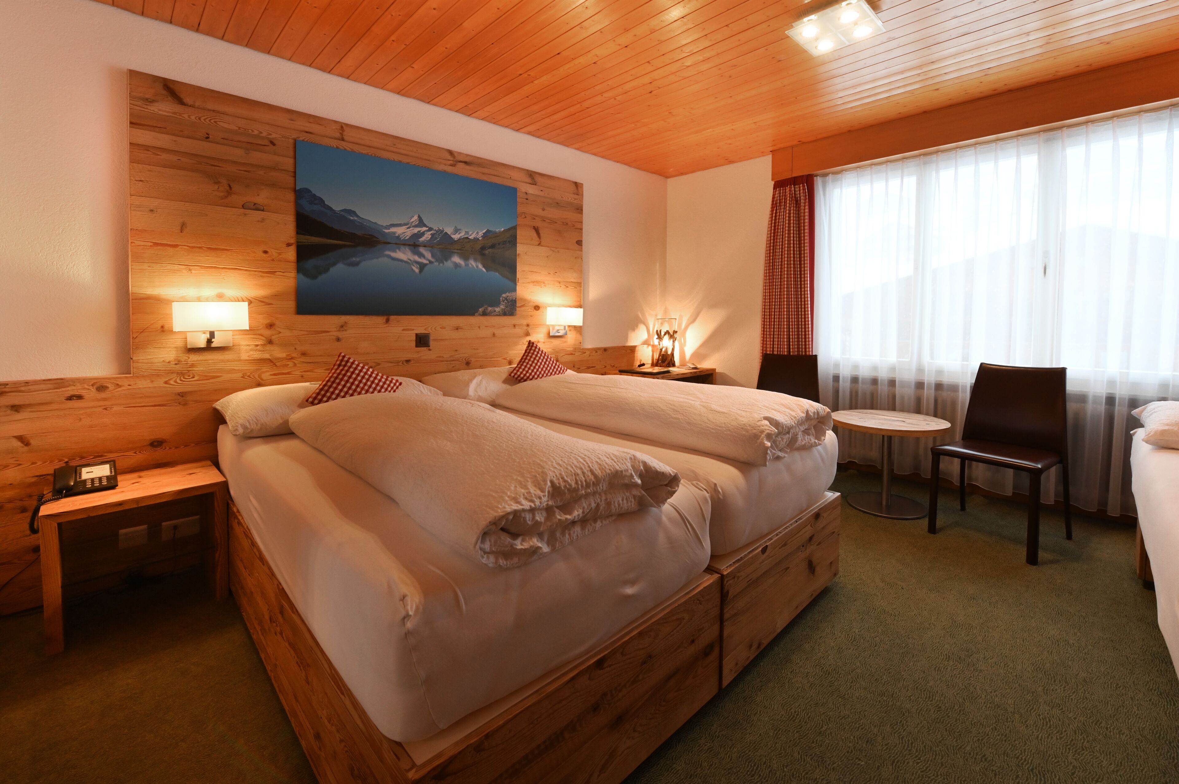 A wooden-paneled room with a large bed, two bedside tables with lamps, a small round table, and a chair.