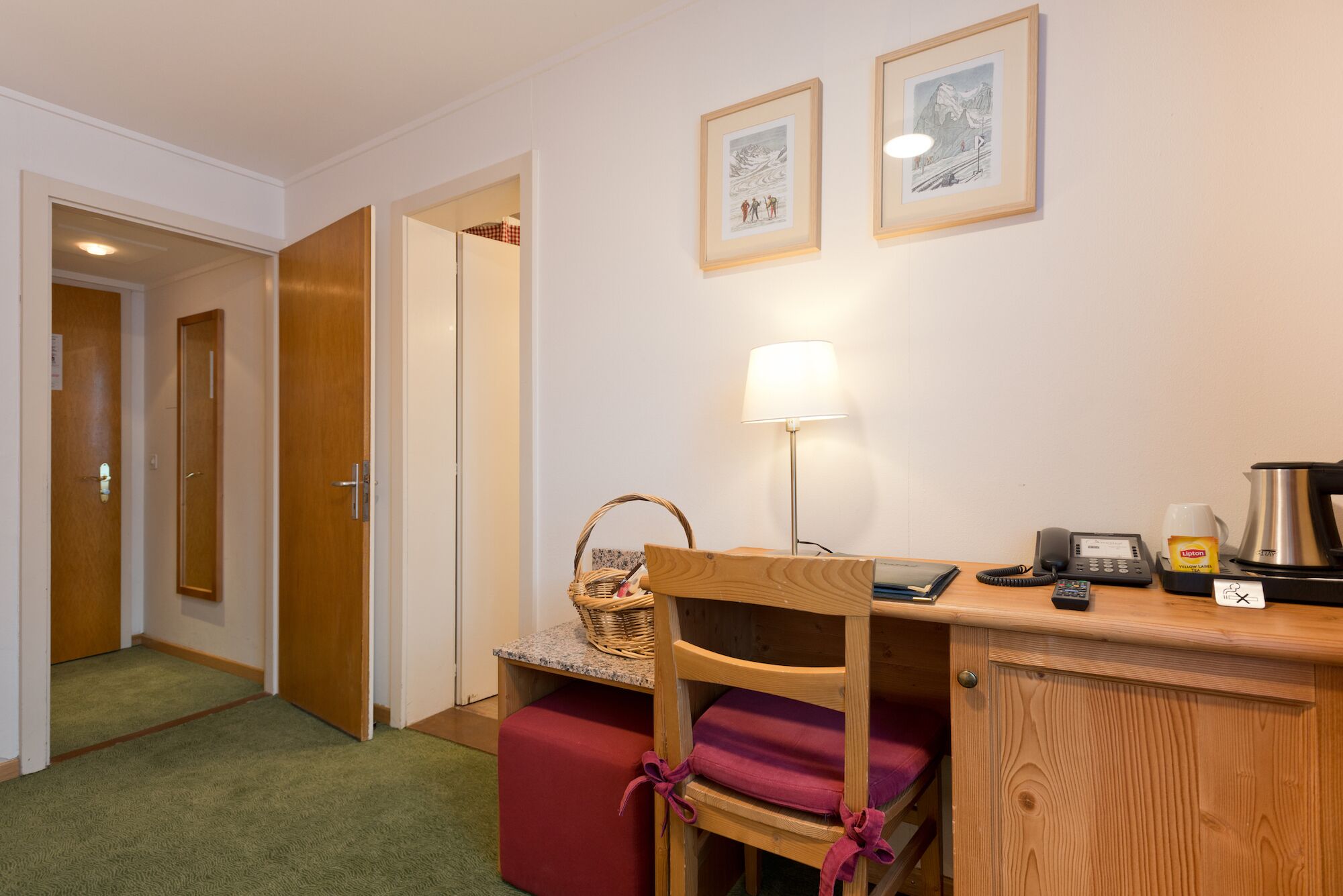A small room with a desk, a chair, a lamp, a phone, and a coffee maker. There are framed pictures on the wall and a door leading to another room.