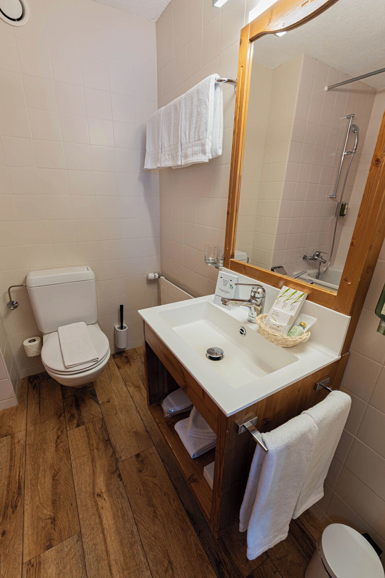 A bathroom with a wooden floor, white tiles, a white toilet, a white sink with a wooden cabinet, a large mirror, and a towel hanging on the wall.