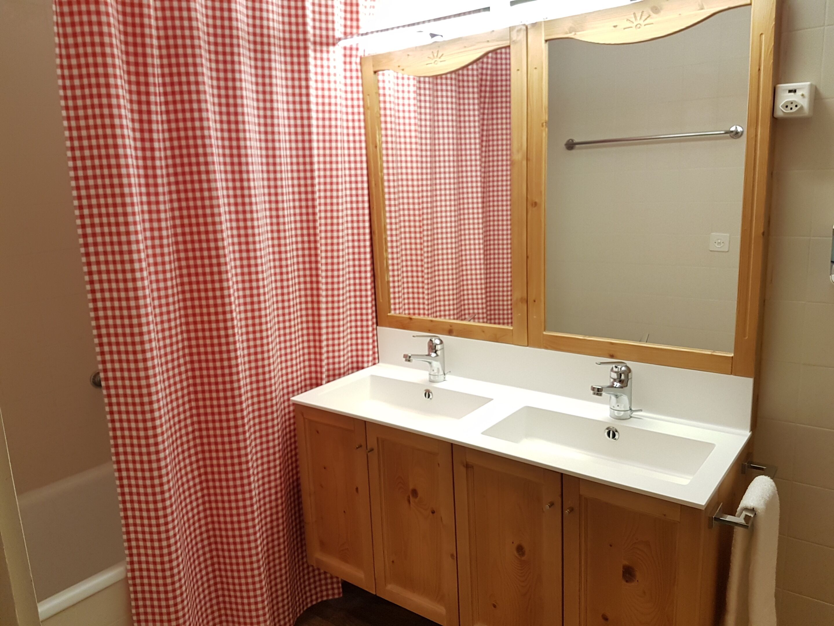 A bathroom with a wooden vanity, two sinks, a mirror, and a red and white checkered shower curtain.
