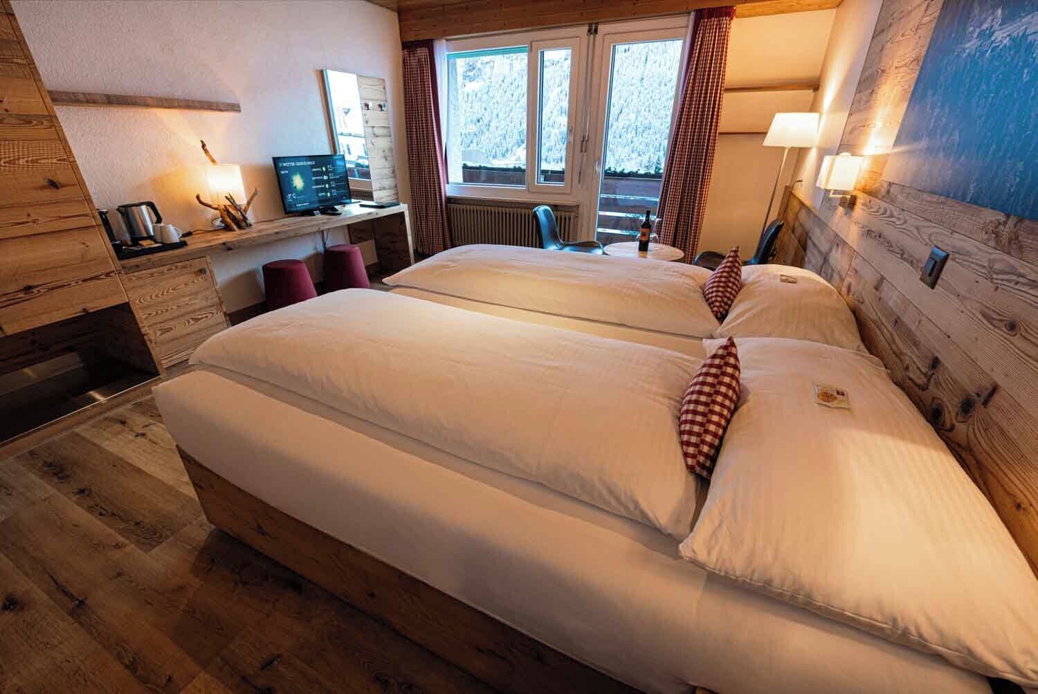 A hotel room with two beds, a desk with a computer, a window with curtains, and wooden walls.