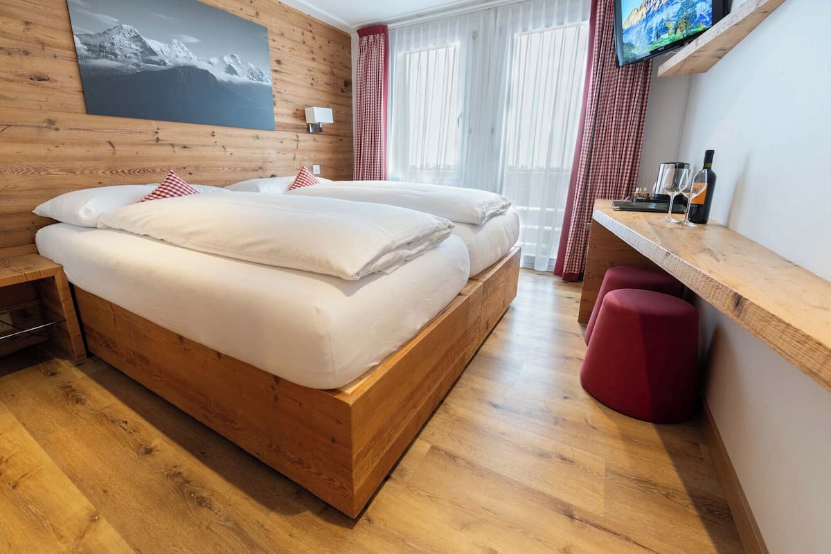 Una cama de madera con ropa de cama blanca, un reposapiés rojo, un escritorio de madera con una botella y vasos, y un televisor en la pared.