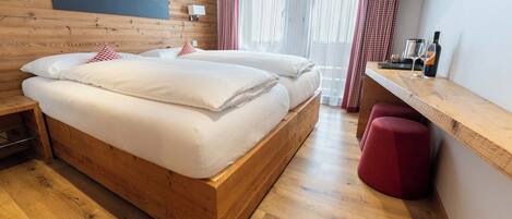 A wooden bed with white bedding, a red ottoman, a wooden desk with a bottle and glasses, and a TV on the wall.