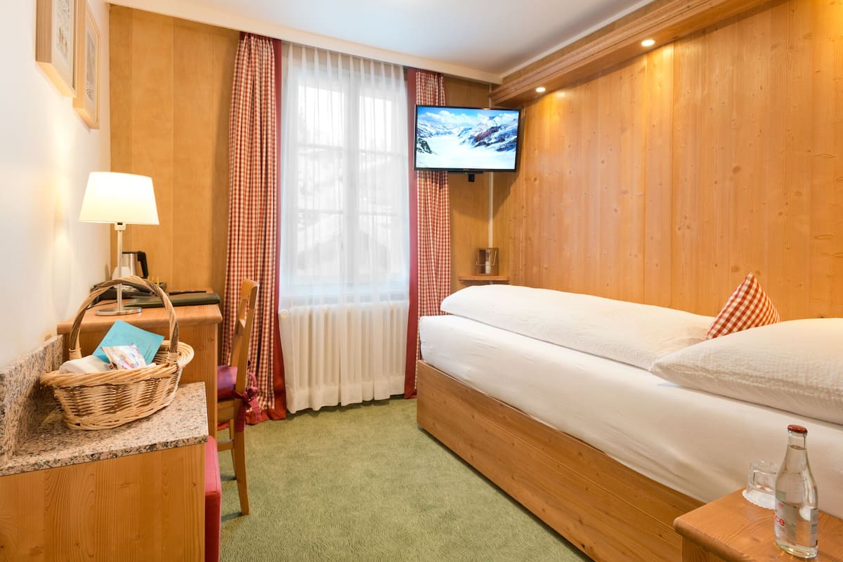 Habitación de hotel con cama de madera, televisor montado en la pared, escritorio con lámpara, cesta de mimbre y botella de agua.