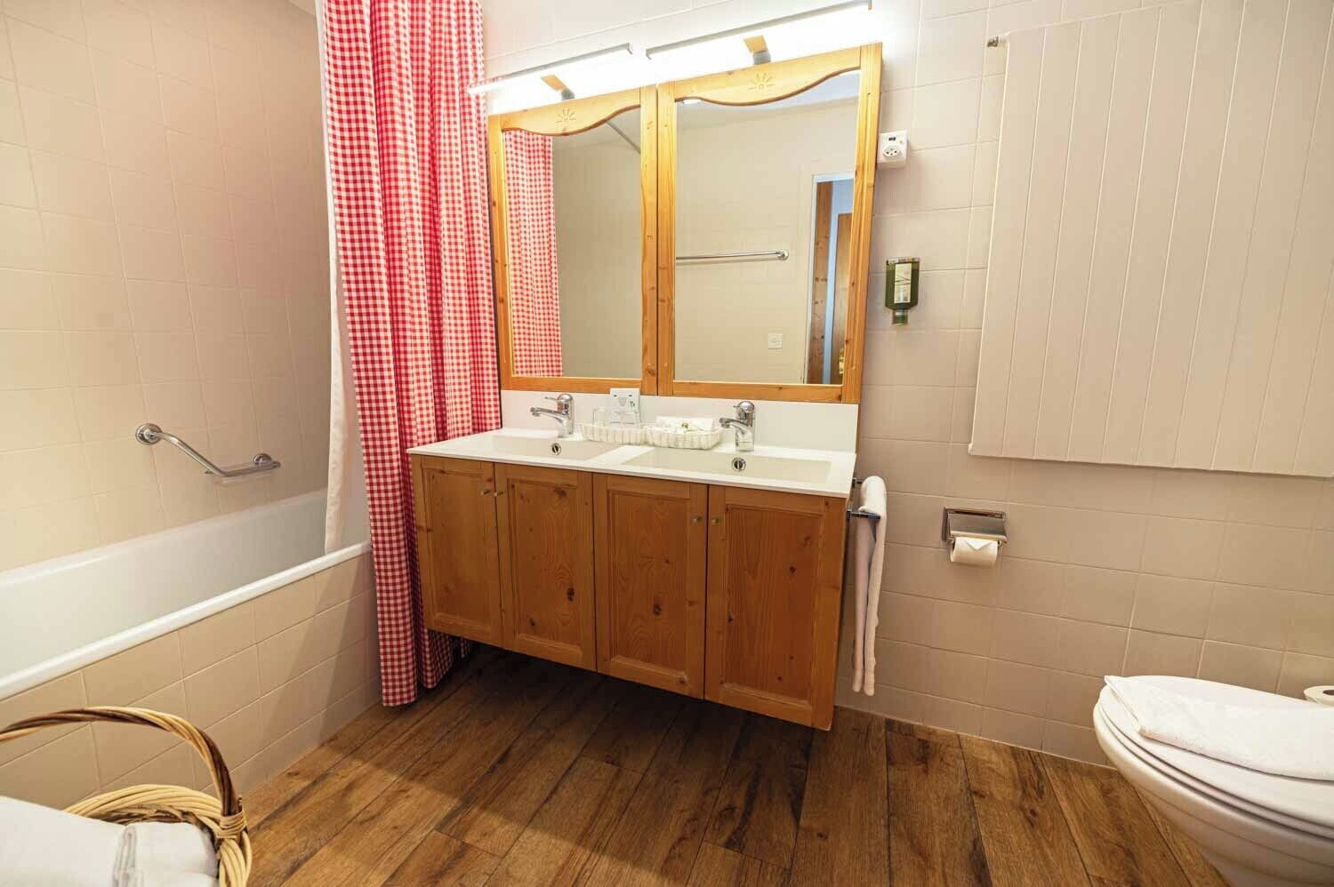 Een badkamer met een houten wastafel, een witte wastafel, een spiegel, een douchegordijn rood-wit geblokt, een houten vloer en een wit toilet.