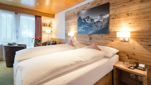 A hotel room with a wooden ceiling, a large bed with white bedding, two bedside lamps, a mounted picture of mountains, and a small bedside table with a phone and a bottle. - Hotel Bernerhof Grindelwald (Grindelwald)