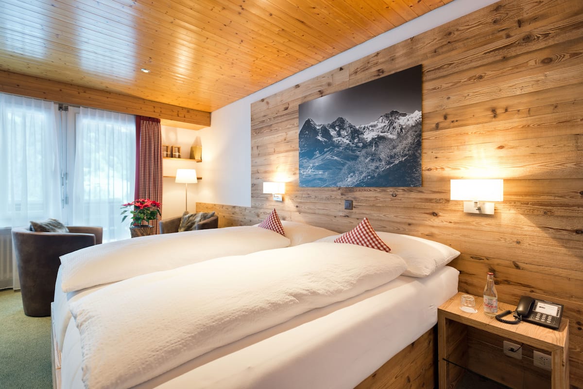 Habitación de hotel con techo de madera, una cama grande con ropa blanca, dos lámparas de noche, un cuadro colgado con paisaje de montañas y una mesita de noche pequeña con un teléfono y una botella.