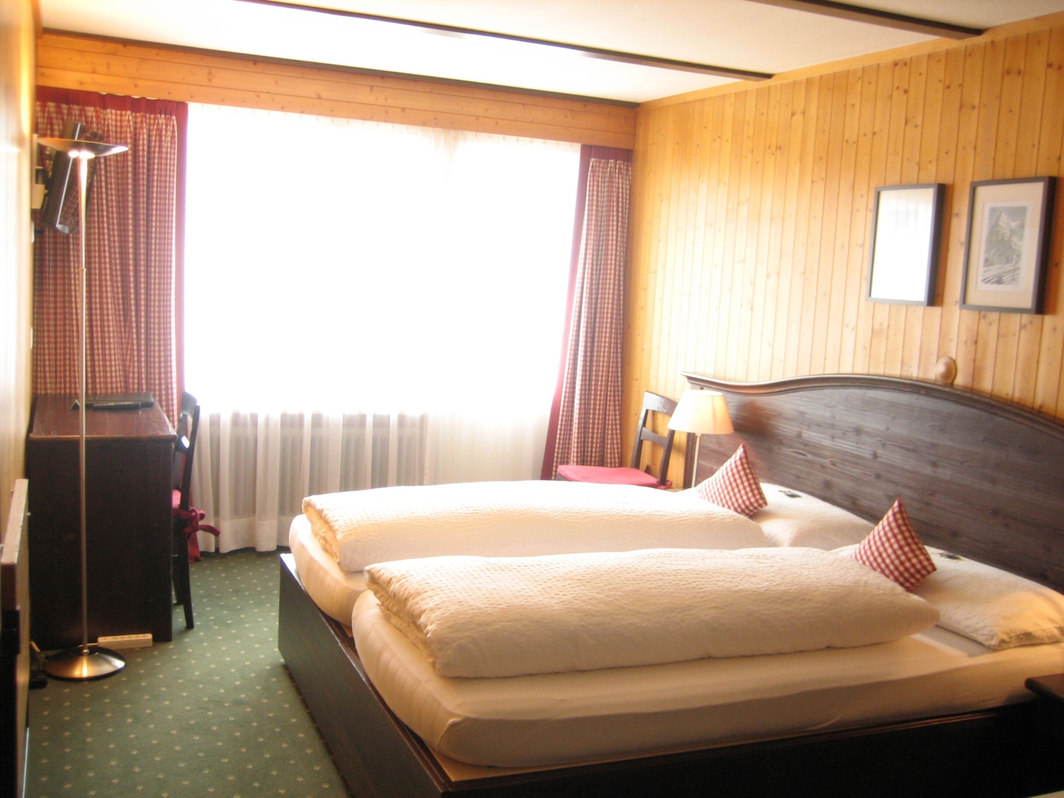 A double bed with two pillows, a wooden headboard, a desk with a lamp, and a window with curtains.