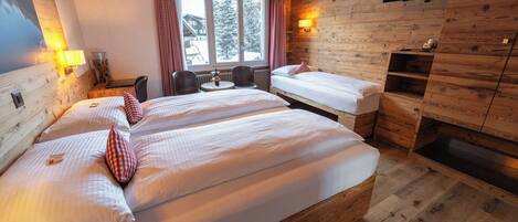 A room with two beds, a TV mounted on the wall, a window with curtains, and a wooden interior.