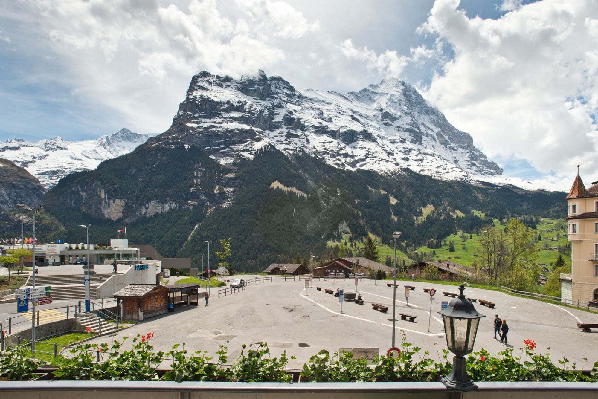 A parking area with a mountain backdrop, a building with a sign reading "PARKING", and a pedestrian pathway leading to a wooden structure.