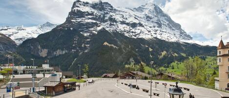 A parking area with a mountain backdrop, a building with a sign reading "PARKING", and a pedestrian pathway leading to a wooden structure.