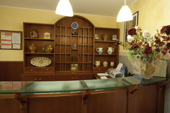 A reception area with a wooden reception desk, a glass counter, and a wall-mounted display of various ceramic items. - Hotel Columbia (Palermo)