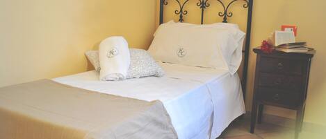 A neatly made bed with white linens and a beige bedspread, a bedside table with a lamp, and a wall-mounted light fixture.