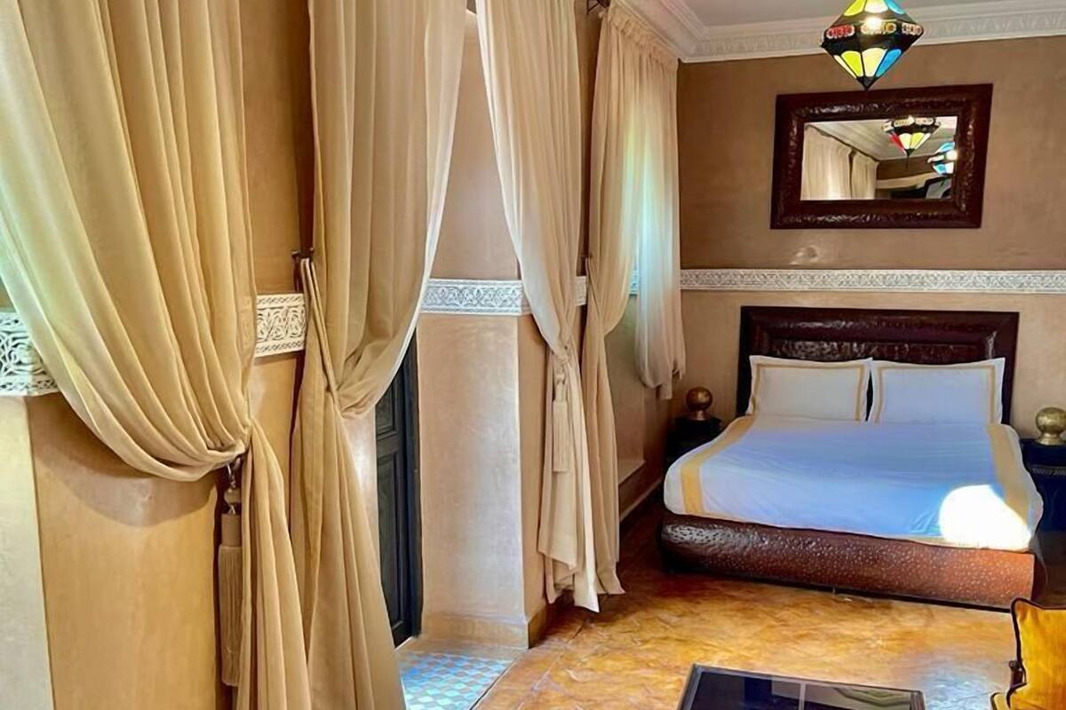 Una habitación de hotel con cama, cortinas, un espejo, una araña y una mesita.