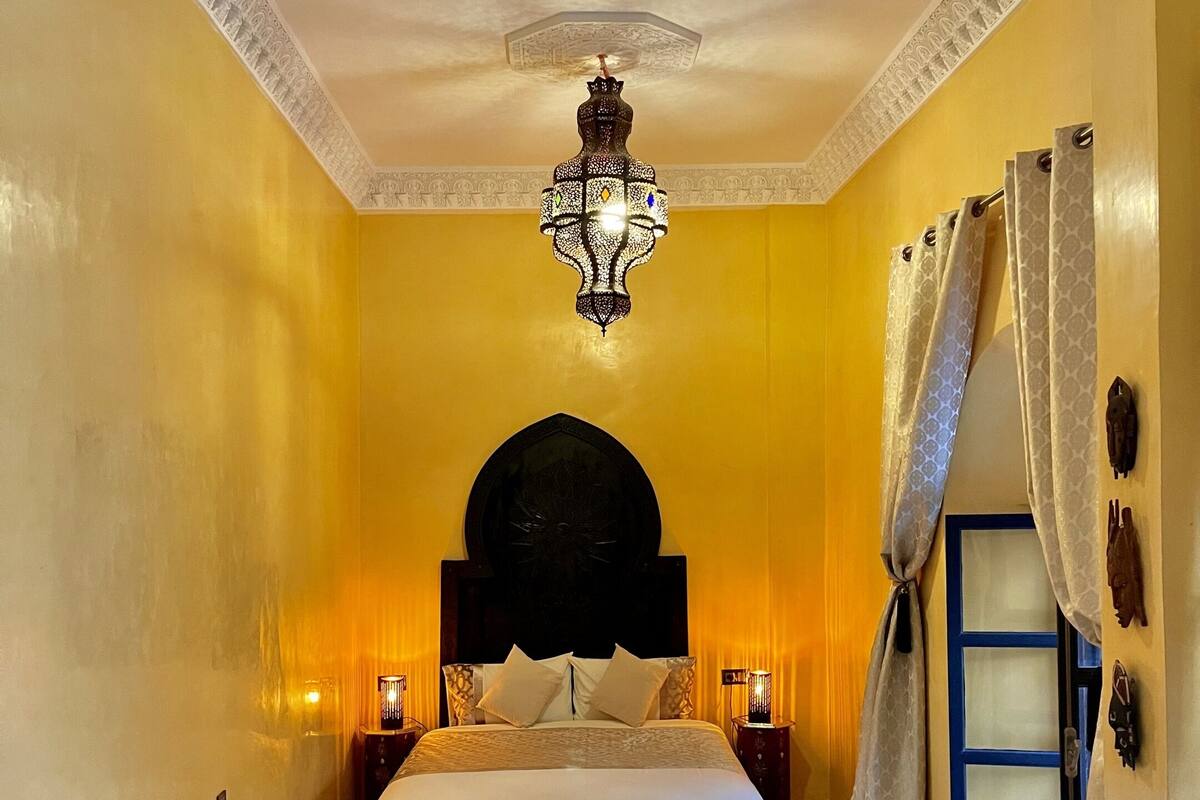 Una habitación con cama, dos sillas, una mesita, una lámpara decorativa en el techo y una ventana con cortinas.