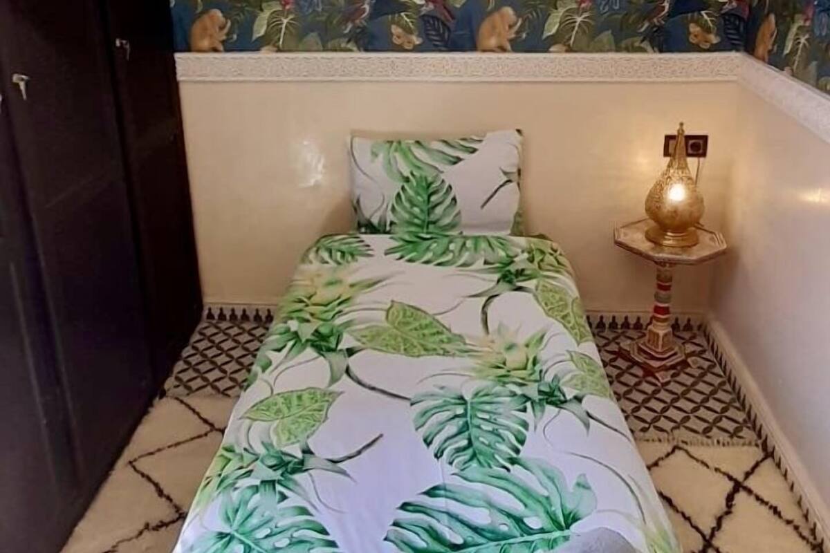 Una habitación con una cama que presenta un diseño de hojas tropicales, un papel tapiz estampado, una lámpara decorativa colgante y una pequeña mesa con lámpara.