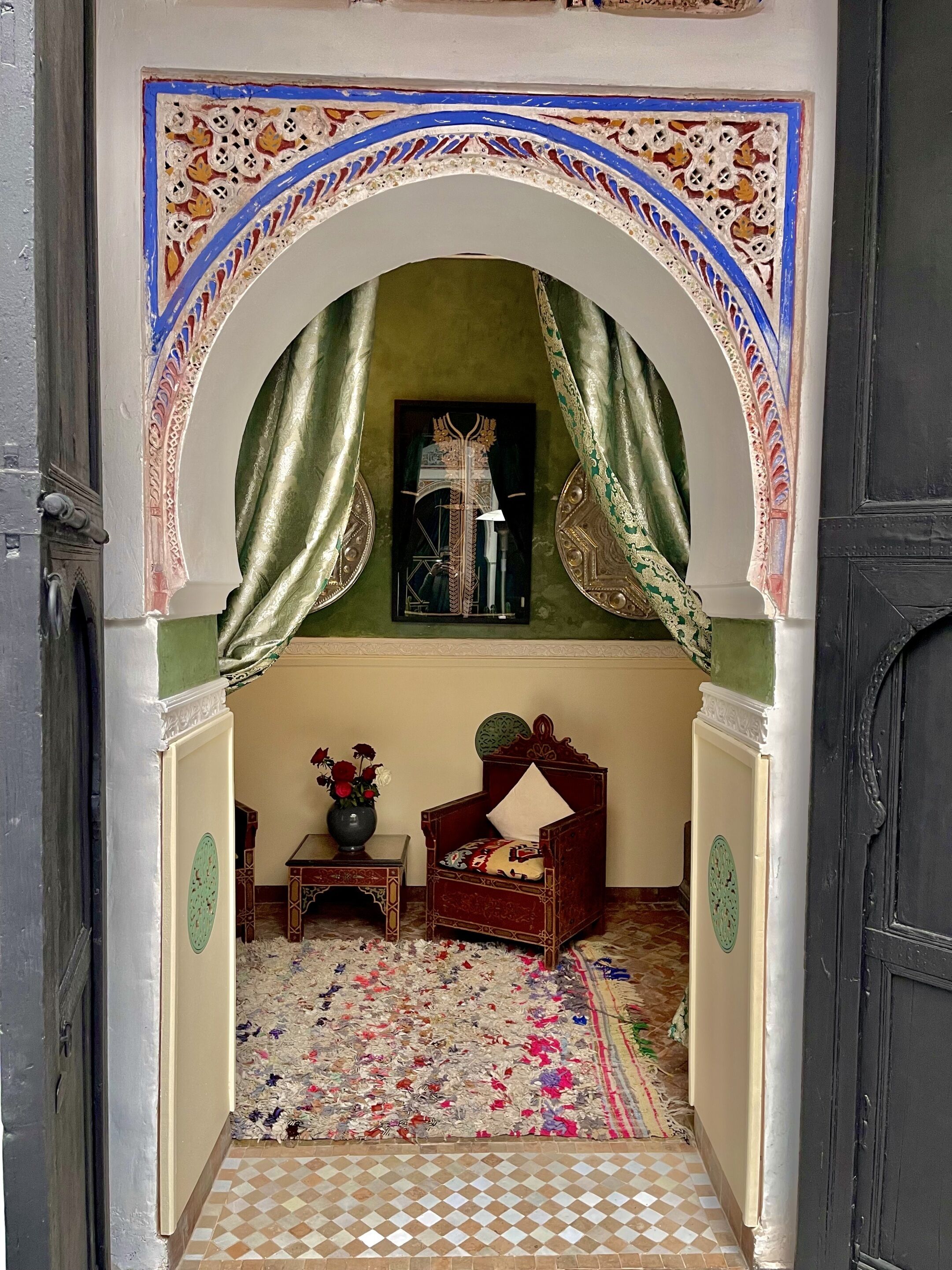 An ornate doorway with a green archway, a bed with a patterned headboard, a small table with a vase of flowers, and a tiled floor.