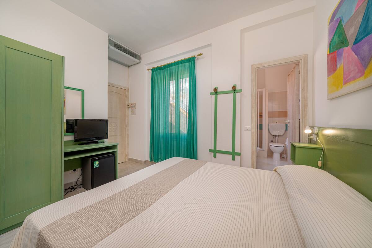 A hotel room with a bed, a television on a desk, a bathroom with a toilet and sink, and a green door.