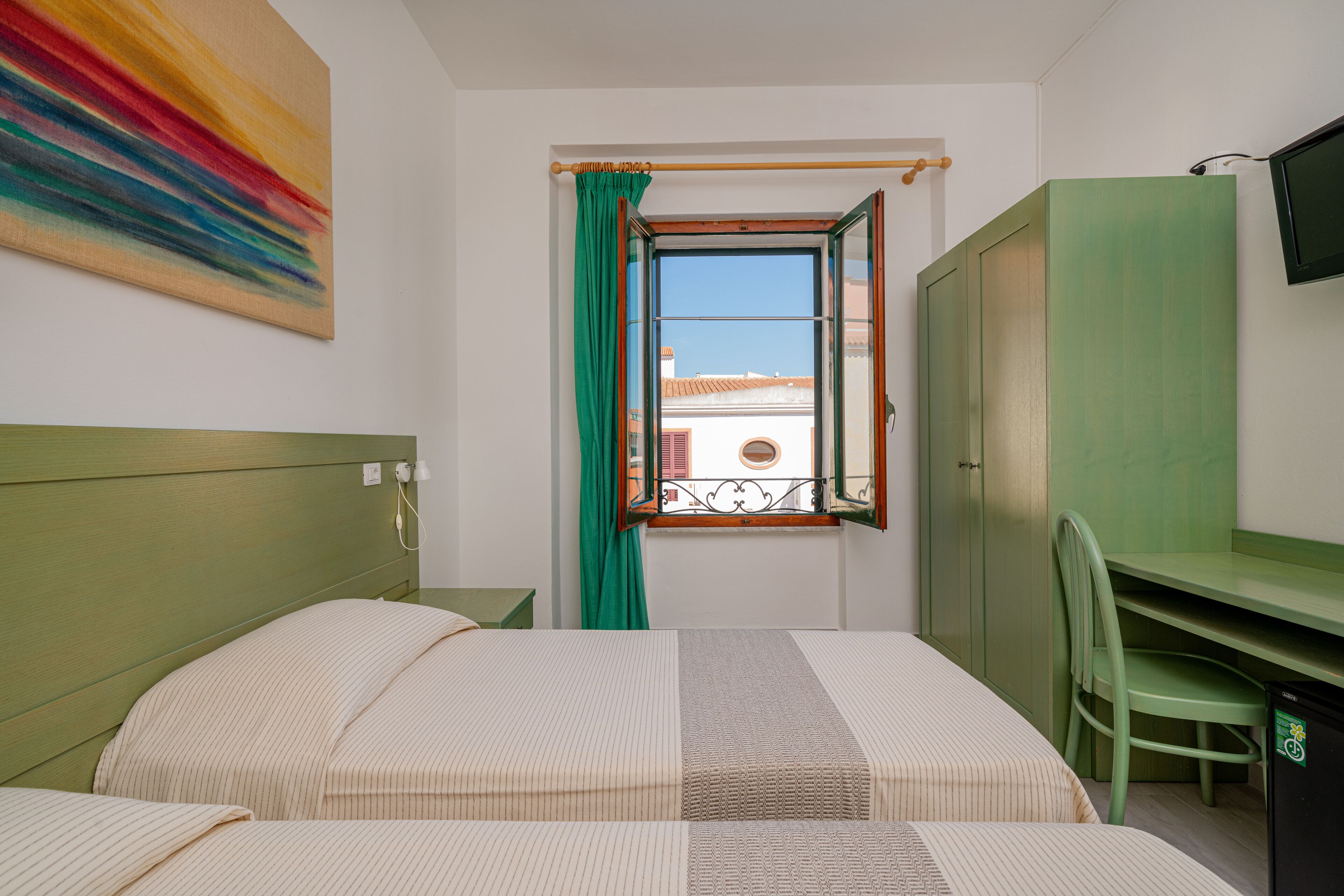 A bedroom with a bed, a desk, and a chair. There is a window with a curtain, a green wardrobe, and a TV mounted on the wall.