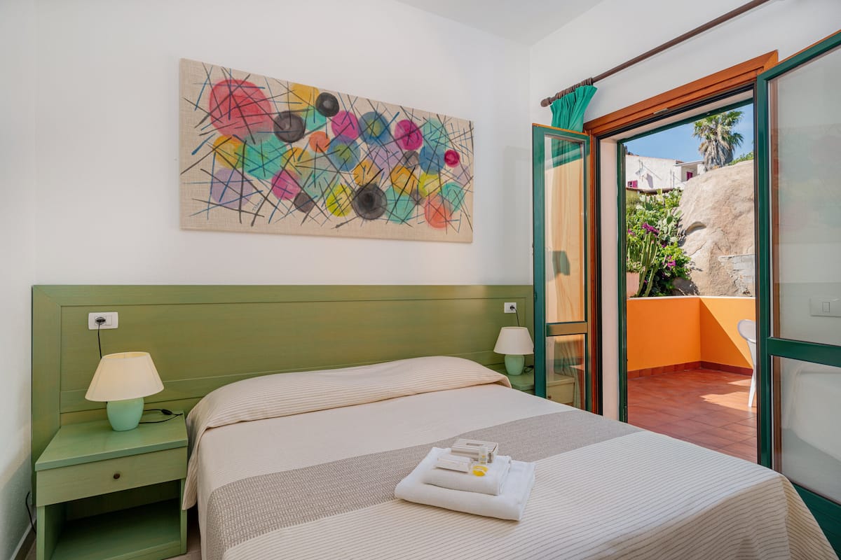 A hotel room with a bed, a nightstand with a lamp, a colorful abstract painting on the wall, and a view of the outside balcony.