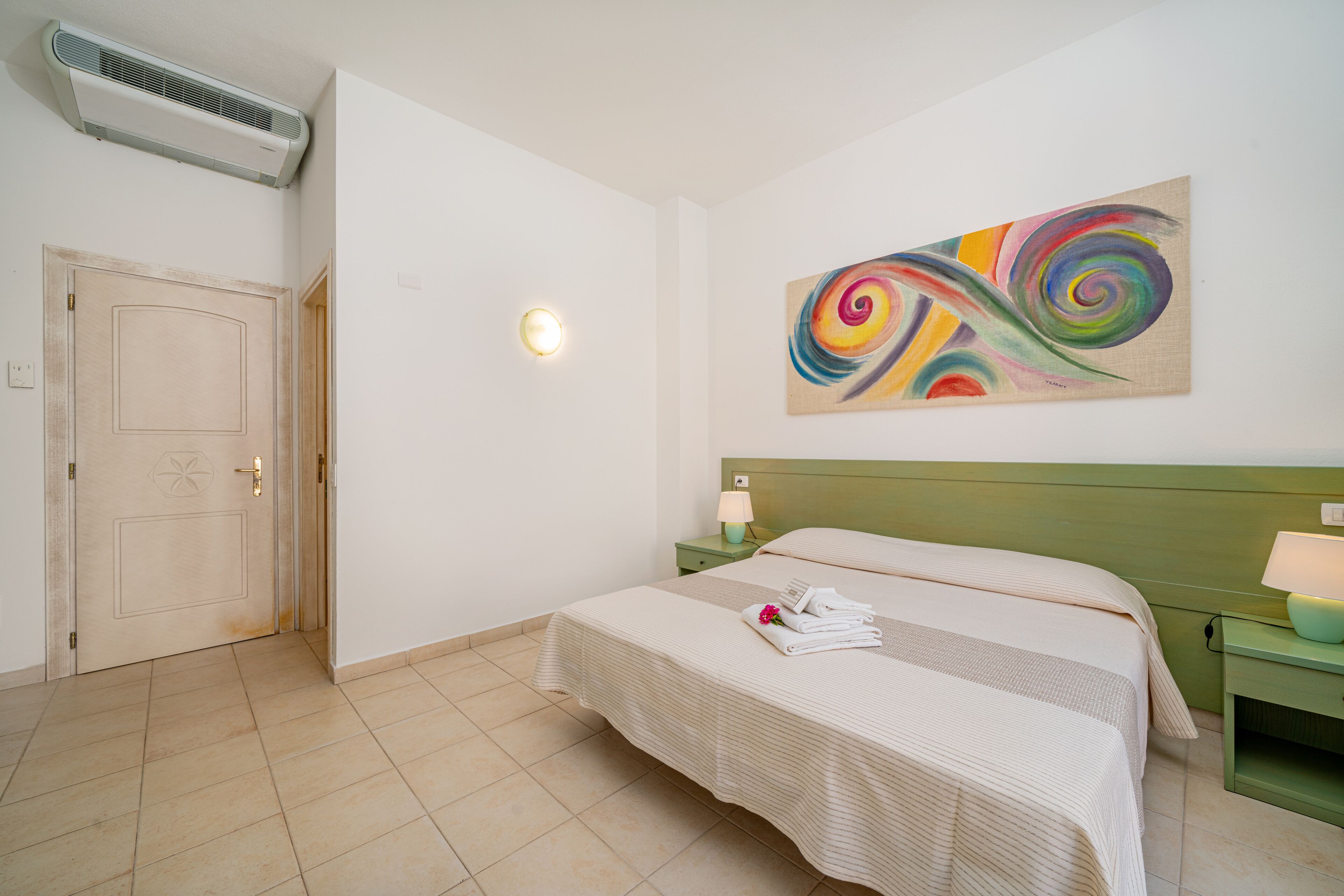 A hotel room with a bed, a green nightstand, a wall-mounted air conditioner, and a colorful abstract painting.