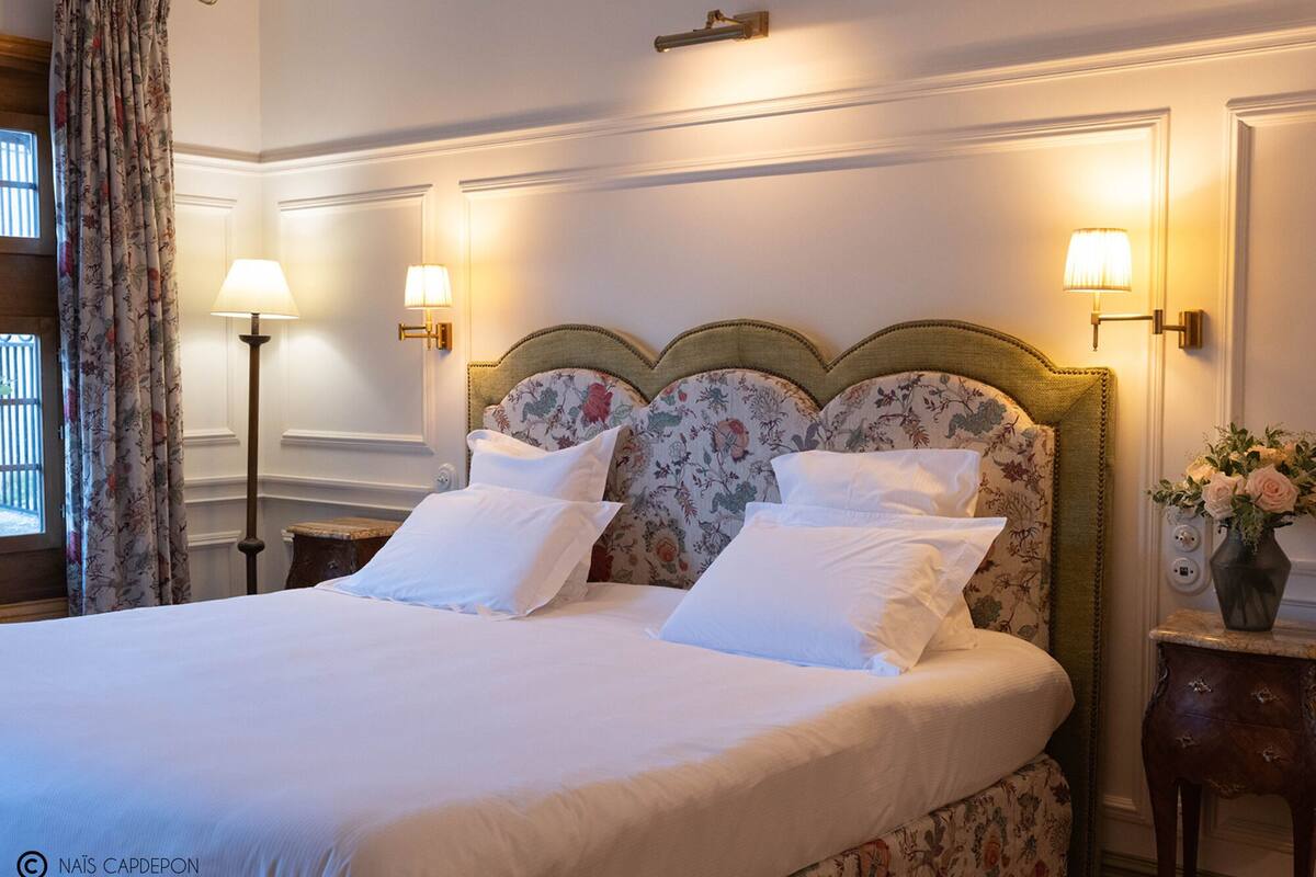 Una cama bien hecha con cabecera floral y almohadas a juego, dos lámparas de noche y un jarrón con flores sobre una mesita.