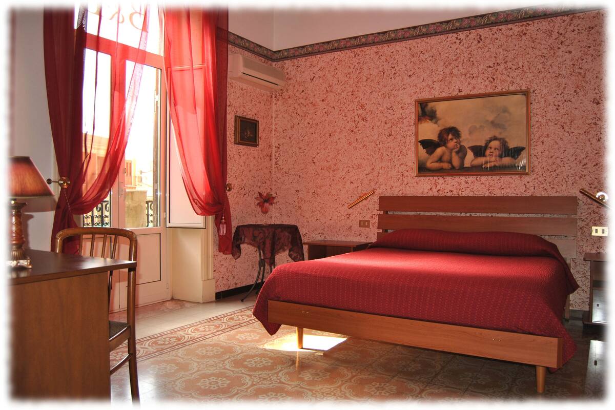 A bedroom with a wooden bed, a desk, and a chair. There is a window with red curtains and a painting on the wall.