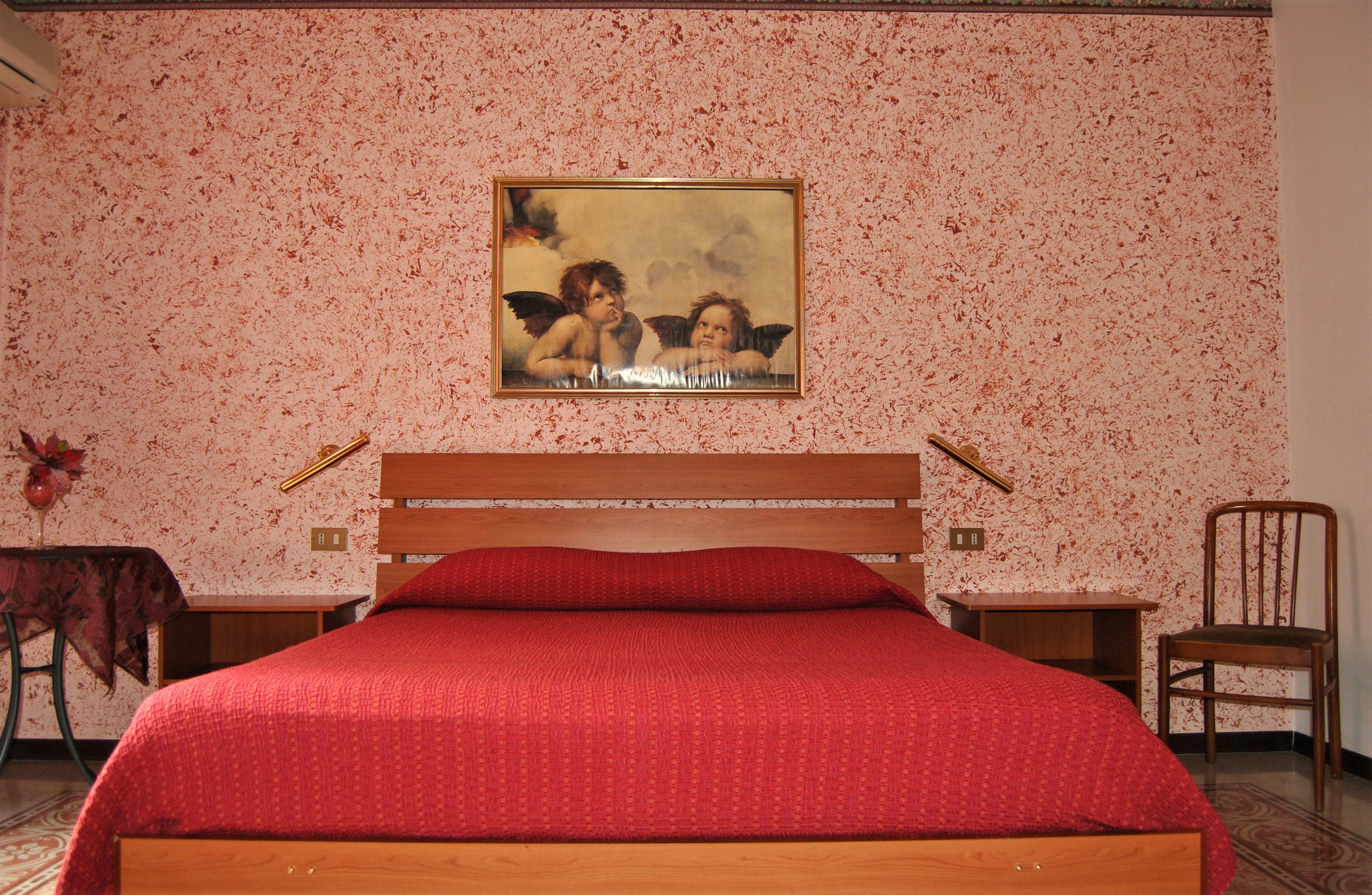 A bedroom with a wooden bed, a red bedspread, a painting of cherubs on the wall, and a small table with a chair.