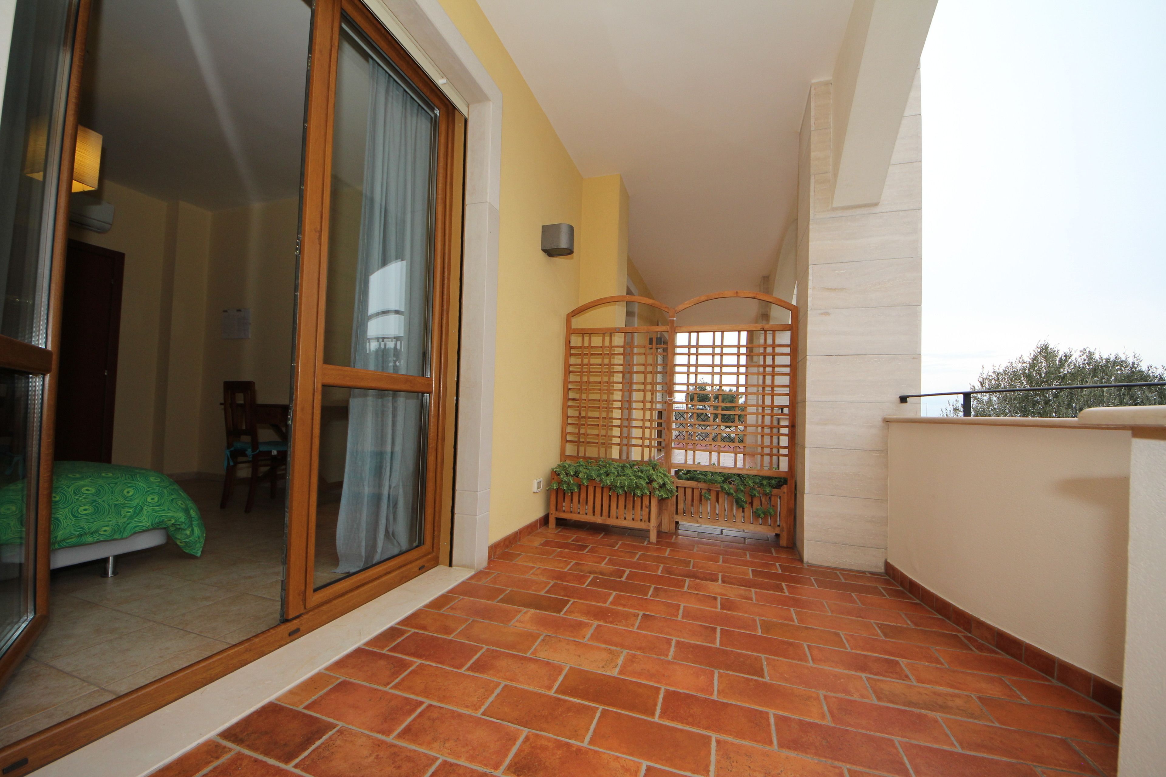 Un balcone con piastrelle in terracotta, una porta scorrevole in vetro che dà su una camera da letto con copripiumino a motivi verdi, e un recinto in legno con vegetazione.