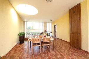 A spacious room with a tiled floor, a dining table with chairs, a large wooden door, and a view through a window.