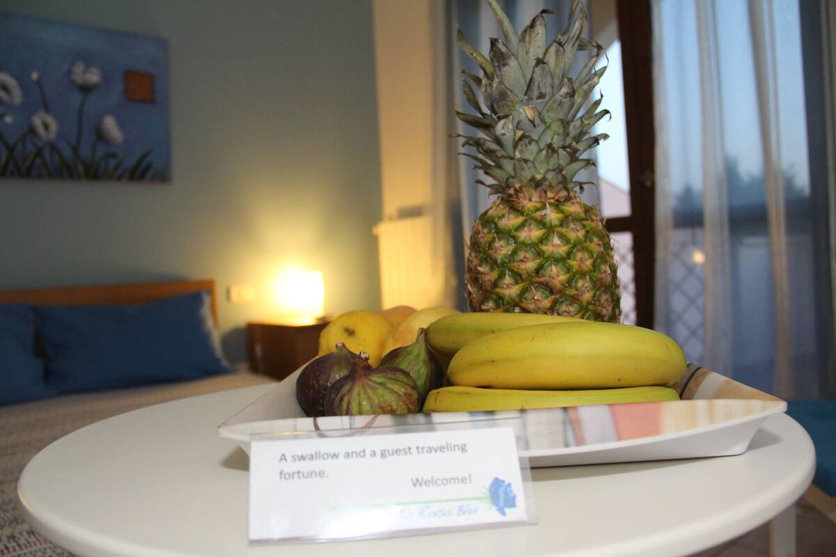 Un panier de fruits contenant des bananes, un ananas et des figues, disposés sur une assiette blanche, avec une pancarte indiquant : « Une hirondelle et un invité, présage de bonne fortune. »