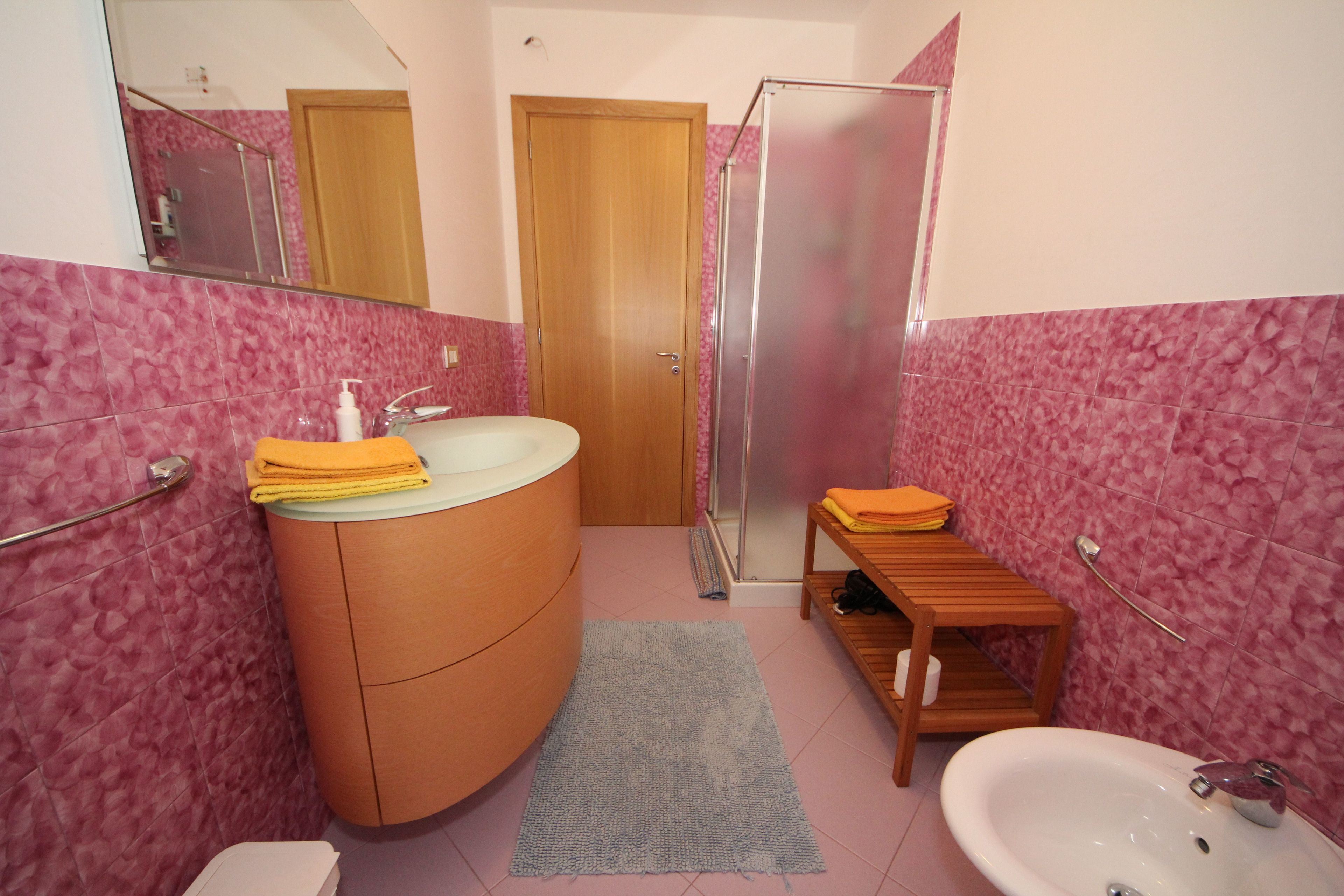 Ein Badezimmer mit rosa gefliesten Wänden, einem Holzwaschtisch, einem Waschbecken, einem Spiegel, einer Dusche und einem kleinen Holzregal mit Handtüchern.