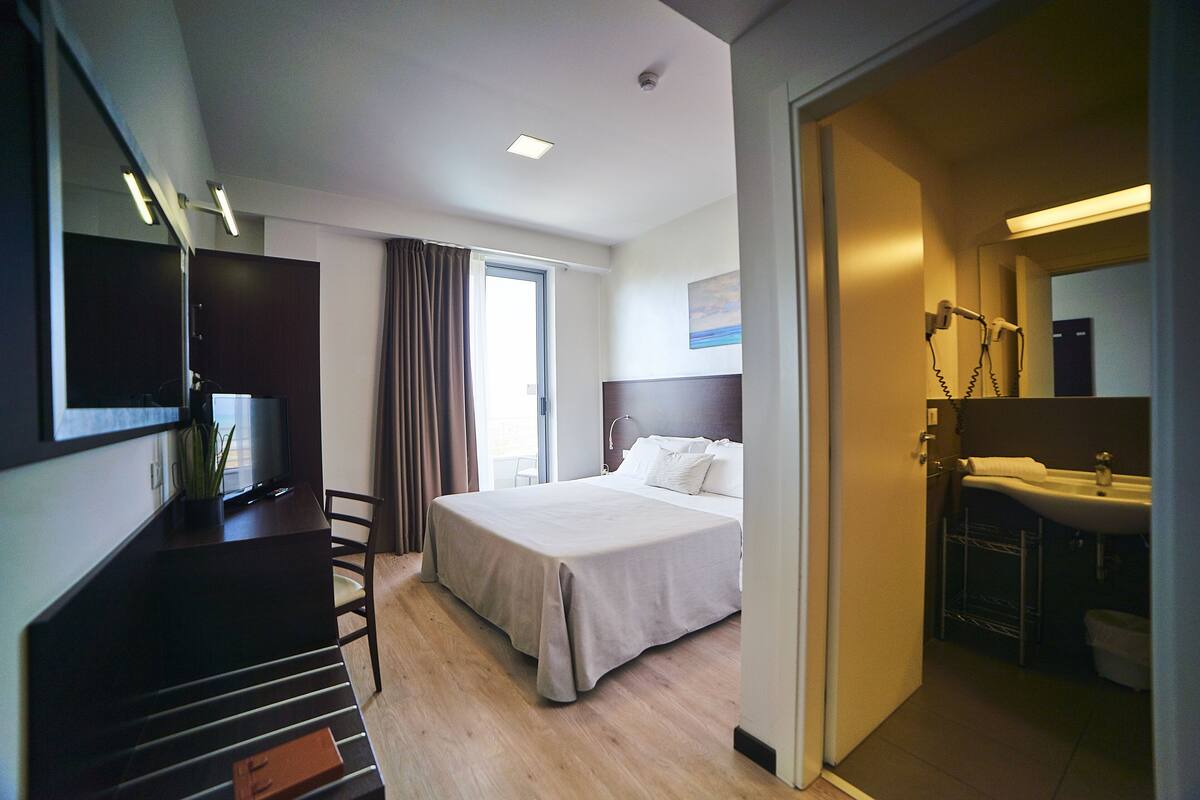 A hotel room with a bed, a desk, a TV, a bathroom with a sink and mirror, and a window with curtains.