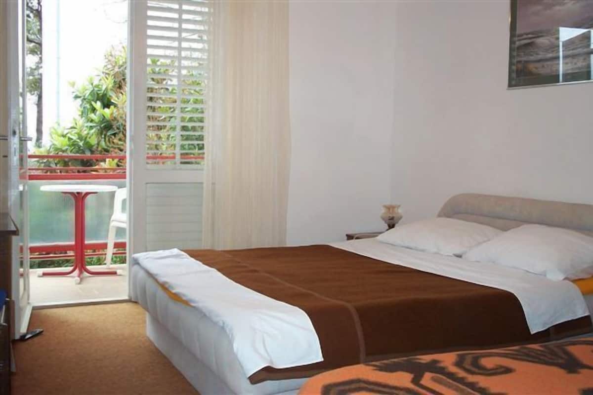Habitación de hotel con cama, mesita de noche, ventana con persianas y vistas a zonas verdes.