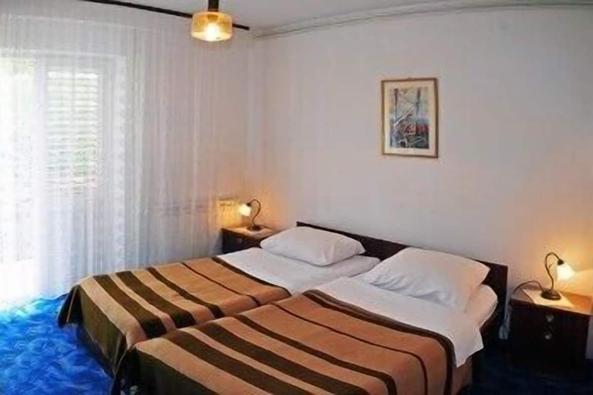Una cama doble con ropa de cama a rayas, una mesita de noche con lámpara, una ventana con cortinas traslúcidas y un cuadro en la pared.