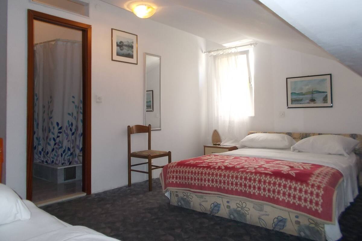 Un dormitorio con cama, una silla, mesita de noche, espejo y ventana con cortinas.