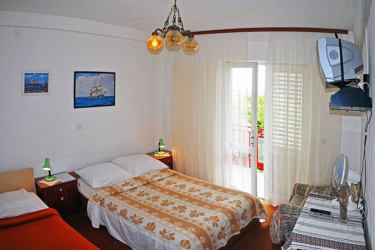 Un dormitorio con una cama, dos mesitas de noche, un escritorio con silla, un ventilador y una ventana con cortinas.