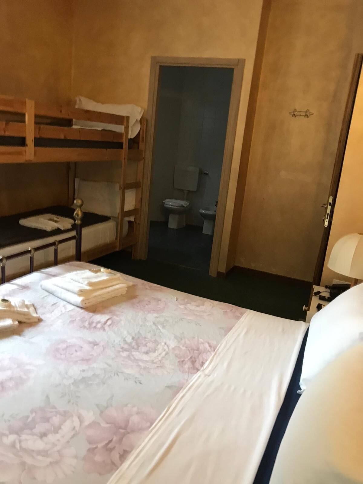 A single bedroom with a bunk bed, a bedside table with a lamp, a floral bedspread, and a door leading to a bathroom.