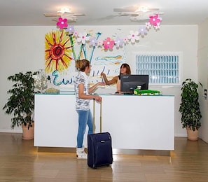 A reception desk with a computer monitor, a plant on each side, and a wall decoration.